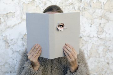 Woman covering face with book, reading poetry, eye looking through cover